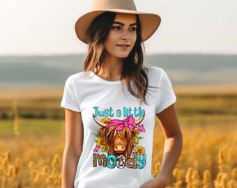 Könnte beinhalten: Weißes T-Shirt mit einer Grafik einer Highland-Kuh, Sonnenblumen und dem Text "Just a little moody". Die Kuh hat eine rosa Schleife und das Wort "moody" ist in einem türkisfarbenen und braunen Muster gehalten. Das T-Shirt wird von einer Person mit einem braunen Hut getragen.