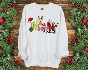 Könnte beinhalten: Weißes Sweatshirt mit dem Text "Nanny Claus" in festlichem Design. Die Buchstaben sind mit Weihnachtselementen wie einem Weihnachtsmannhut, Rentier und Geschenkboxen verziert. Der Hintergrund zeigt Tannenzweige und rote Beeren.