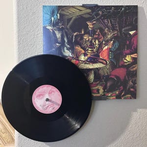 May include: A black vinyl record with a pink label featuring a face illustration. Next to it, a framed print shows a whimsical scene with anthropomorphic animals in a colourful, vintage style, reminiscent of a storybook illustration.