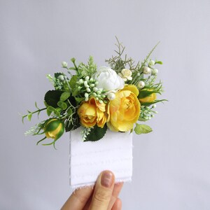 May include: A small bouquet of yellow and white artificial flowers with green leaves. The flowers are arranged on a white card.