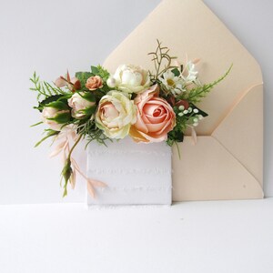 May include: A peach-colored envelope with a bouquet of flowers and a white card. The flowers include peach, cream, and white roses, with green foliage. The card has a textured, fringed edge.