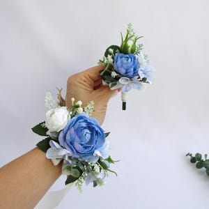 Op de afbeelding: Een blauwe en witte bloemen polscorsage en bijpassende corsage. De corsage heeft een grote blauwe roos, witte rozen en groen. De corsage is kleiner en heeft een blauwe roos en groen.