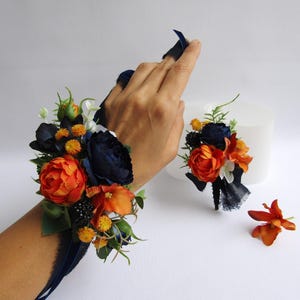 May include: A wrist corsage and a matching boutonniere featuring orange and navy blue flowers with greenery. The corsage is worn on a wrist with a navy blue ribbon. The boutonniere is on a white surface.