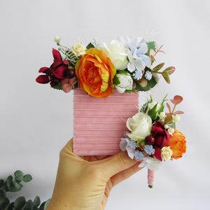 May include: Two floral boutonnieres with a pink velvet box. The boutonnieres feature orange, white, red, and blue flowers.