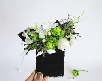 May include: A black rectangular card adorned with a vibrant floral arrangement. The bouquet features white and green flowers, accented by sprigs of greenery and dark accents. The card is held by a hand against a white background.