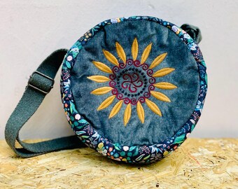 May include: Round denim crossbody bag with a sunflower embroidery in yellow and burgundy. The bag has a floral patterned border and a green adjustable strap. The bag is on a wooden surface.