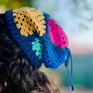 Op de afbeelding: Een kleurrijke gehaakte hoofdbedekking met een patchwork ontwerp. De hoofdbedekking is voornamelijk blauw, met gouden, roze en turquoise secties. Het haakwerk is zichtbaar, met een getextureerde uitstraling. De hoofdbedekking wordt op het hoofd van een persoon gedragen.