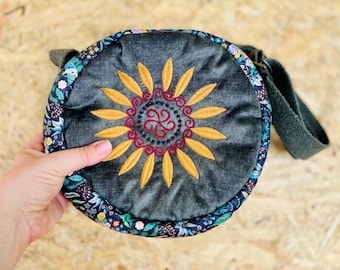 May include: A round, dark green fabric bag with a floral print border. The bag has a long strap and a circular embroidered design in the center featuring a yellow sun with red accents.