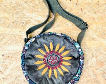 May include: A round, dark green crossbody bag with a floral print border. The bag has a large embroidered sunflower with a red and gold triquetra in the center.