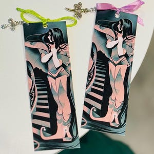 以下が含まれることがあります： 長い黒髪とピンクとティールの流れるようなガウンを着た女性の様式化されたイラストが特徴の2つのブックマーク。1つは緑のリボンと十字架のチャーム、もう1つはピンクのリボンとトンボのチャームが付いています。