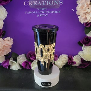 May include: A black tumbler with a gold glitter design that says "Dope" with gold dripping down the side. The tumbler is on a white rotating display stand.