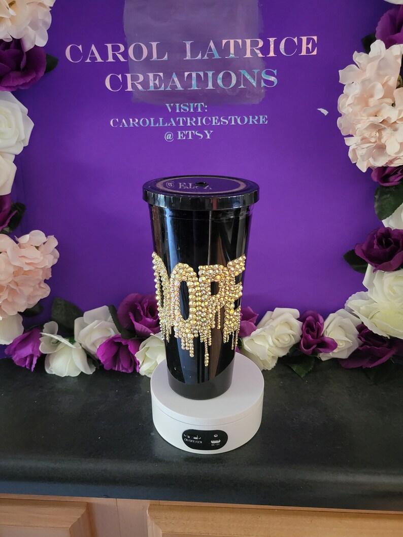 May include: A black tumbler with a gold glitter design that says "Dope" with gold dripping down the side. The tumbler is on a white rotating display stand.