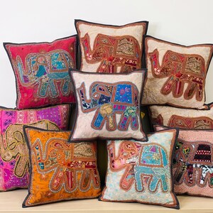 May include: A collection of decorative throw pillows with elephant designs. The pillows feature a variety of colors, including red, pink, orange, and blue, with intricate patchwork and embellishments. Each pillow has a dark border.