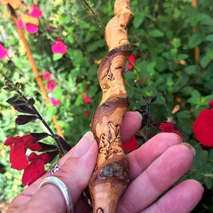 May include: A hand-carved wooden wand featuring a natural, twisted, spiral shape with a smooth, polished finish. The wood displays varied light and dark brown grain patterns, with intricate, dark, etched leaf designs visible along the surface.