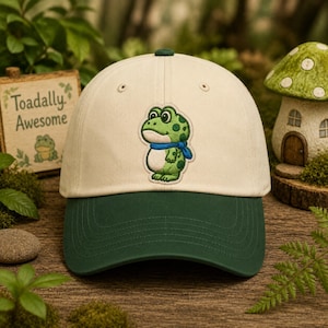 Könnte beinhalten: Eine cremefarbene Baseballkappe mit dunkelgrünem Schirm. Auf der Vorderseite befindet sich eine Stickerei eines grünen Frosches mit blauem Schal. Links steht ein Holzschild mit dem Text "Toadally Awesome" über einer kleinen Froschabbildung.