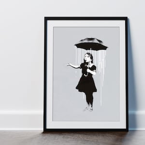 May include: A black and white framed print of a girl standing under a black umbrella with white rain streaks. The girl is wearing a black dress and holding the umbrella handle.