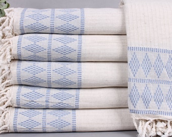 May include: A stack of off-white cotton towels with blue geometric patterns and fringed edges. The towels are folded neatly, showcasing the detailed diamond and square designs. The soft color palette and texture suggest a spa or bathroom theme.