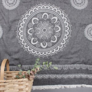 May include: Gray beach towel with white mandala pattern. The towel has a large central mandala design and smaller circular patterns. A decorative border and fringe complete the design. A basket with greenery is in the foreground.