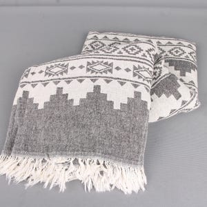 Pode incluir: Uma manta dobrada com um padr&atilde;o geom&eacute;trico cinza e branco. A manta tem uma borda com franjas. O padr&atilde;o inclui formas de diamante e tri&acirc;ngulo. A manta &eacute; feita de um material macio.