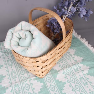 Pode incluir: Uma cesta de vime com uma al&ccedil;a cont&eacute;m uma toalha enrolada com padr&atilde;o azul claro e branco. A cesta est&aacute; sobre uma toalha estampada combinando com borda de franjas. Um raminho de flores roxas est&aacute; na cesta.