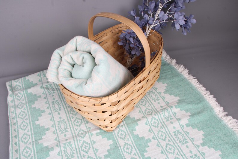 Pode incluir: Uma cesta de vime com uma al&ccedil;a cont&eacute;m uma toalha enrolada com padr&atilde;o azul claro e branco. A cesta est&aacute; sobre uma toalha estampada combinando com borda de franjas. Um raminho de flores roxas est&aacute; na cesta.