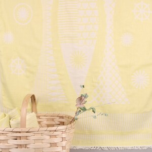 May include: A yellow beach towel with a white geometric design featuring sun and ship wheel motifs. Rolled towels are in a woven basket with a handle. The towel has a fringed edge. A sprig of greenery is placed near the basket.