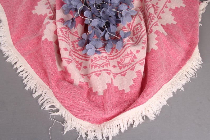Pode incluir: Uma toalha de praia redonda com estampa rosa e branca com borda com franjas. A toalha apresenta um design geom&eacute;trico e &eacute; adornada com raminhos decorativos azuis.