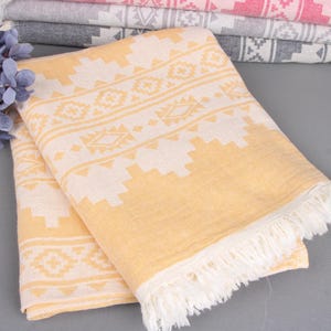 Pode incluir: Uma toalha dobrada com padr&atilde;o amarelo e branco com borda com franjas. A toalha tem um design geom&eacute;trico e faz parte de uma pilha de toalhas semelhantes em v&aacute;rias cores. Flores azuis decorativas no fundo.