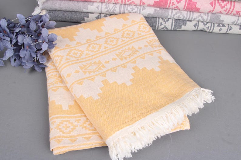 Pode incluir: Uma toalha dobrada com padr&atilde;o amarelo e branco com borda com franjas. A toalha tem um design geom&eacute;trico e faz parte de uma pilha de toalhas semelhantes em v&aacute;rias cores. Flores azuis decorativas no fundo.