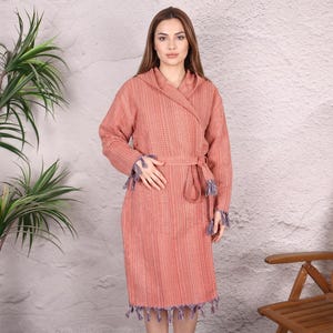 May include: A coral-colored, long-sleeved robe with a wrap-around design and a matching belt. The robe features fringed edges and cuffs with blue and purple accents. A wooden chair and a plant are in the background.