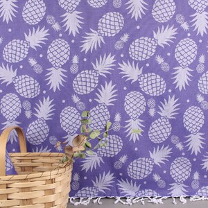 Puede incluir: Una toalla de playa de color morado con un patrón de piña blanco. La toalla tiene flecos en el borde inferior.