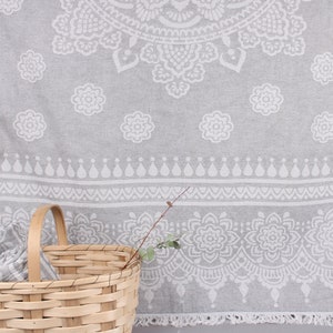May include: A gray and white patterned beach towel with a floral design, displayed with a wicker basket. The towel has a fringe along the bottom edge and is folded inside the basket.