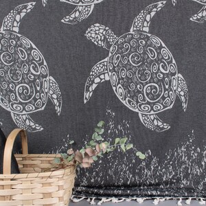 May include: Dark gray beach towel with a white turtle pattern. The towel is folded and placed in a light brown wicker basket. The towel has a fringe along the bottom edge. A sprig of greenery is placed in front of the towel.