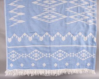 May include: A light blue and white patterned beach towel with a geometric design. The towel has a fringed edge and is made of a soft, woven material. The pattern includes diamond and triangle shapes.