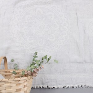 May include: A light gray linen textile with a large, circular, floral design. A woven basket with a handle and a sprig of greenery are in the foreground. The textile has a fringed edge.