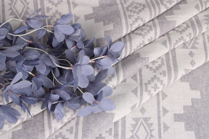 Pode incluir: Um tecido cinza e branco dobrado com um padr&atilde;o geom&eacute;trico. Um ramo de flores azuis artificiais repousa sobre o tecido. O tecido &eacute; dobrado para mostrar o padr&atilde;o e a textura.