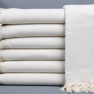 Puede incluir: Una pila de seis mantas crema con estampado de espiga y bordes con flecos. Las mantas están cuidadosamente dobladas, mostrando el tejido texturizado. El color neutro y el diseño sencillo sugieren comodidad y estilo.