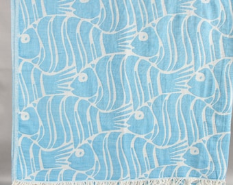 May include: A light blue and white patterned beach towel. The towel features a repeating pattern of stylized fish. The fish are white and outlined with a light blue background. The towel has a fringe on the bottom edge.