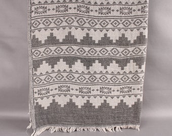 May include: A gray and white patterned textile with a geometric design. The fabric features repeating patterns of diamonds, triangles, and other shapes. The edges are finished with a fringe. This textile could be a blanket, throw, or decorative cloth.