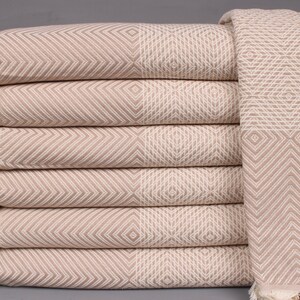 May include: A stack of beige and white patterned Turkish towels with fringed edges. The towels feature a geometric design of diamonds and chevrons. The towels are folded neatly, showcasing the intricate weave and texture of the fabric.