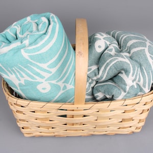 May include: A wicker basket contains two rolled beach towels. One is turquoise with a white leaf pattern, the other teal with a similar white design. The basket has a wooden handle and is set against a grey background.