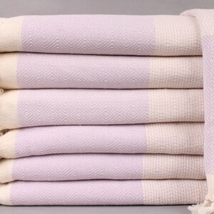 May include: Stack of six lavender and cream-colored Turkish towels. Each towel features a diamond pattern in the lavender section and cream-colored fringe. The towels are neatly folded and stacked, showcasing their texture and design.