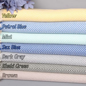 May include: A stack of seven cotton towels in various colors, including yellow, petrol blue, mint, sax blue, dark gray, khaki green, and brown. Each towel has a herringbone pattern.