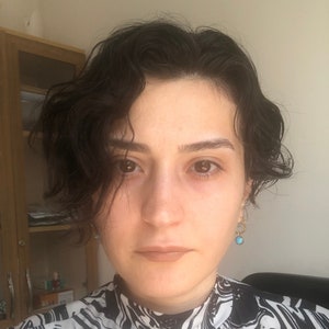 May include: A person with dark wavy hair and brown eyes. They are wearing a black and white patterned top and small turquoise earrings. The person is looking directly at the camera.