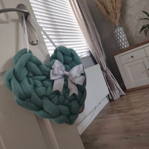 May include: A teal-coloured, chunky knit heart decoration with a white satin bow, suspended by a white ribbon. The heart is made of thick yarn and is a handmade item. The decoration is a cozy item, perfect for home decor.