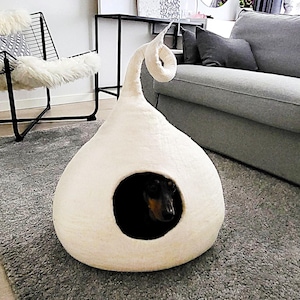 Puede incluir: Una cueva para mascotas de fieltro color crema, con forma de gota, punta curvada y una abertura frontal circular. Un perro pequeño de color negro y fuego se encuentra dentro de la estructura, situada sobre una alfombra gris.