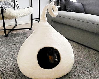 May include: A cream-colored, teardrop-shaped felt pet cave with a pointed, curled top and a circular front opening. A small black and tan dog is visible inside the structure, which is resting on a gray rug in a living room setting.