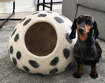 May include: A black and tan dachshund stands next to a round, off-white felt pet cave featuring dark gray circular spots. The pet cave has a large, circular opening on the front, providing a cozy, enclosed space for a small animal to rest inside.