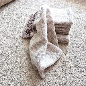 May include: A stack of folded beige blankets with a matching throw blanket with fringe. The blankets are on a light beige carpet, with a beige sectional sofa in the background.