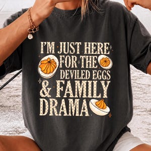 May include: A charcoal gray short-sleeve t-shirt featuring the text "I'M JUST HERE FOR THE DEVILED EGGS & FAMILY DRAMA" in a cream-colored, dotted font, accompanied by illustrations of three halved deviled eggs with orange yolks.